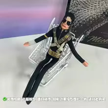 A Michael Jackson doll dressed in a black and gold jacket and pants, reclining on a clear plastic chair against a white and purple background.